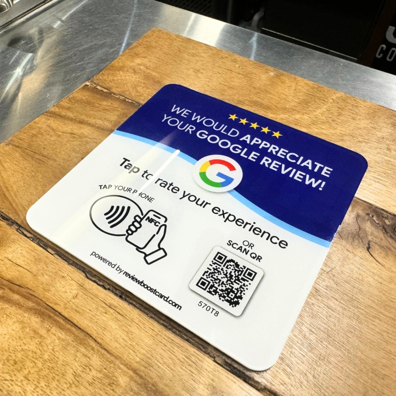 A ReviewBoost plaque on a wooden counter, encouraging customers to leave a Google review by tapping their phone or scanning the QR code