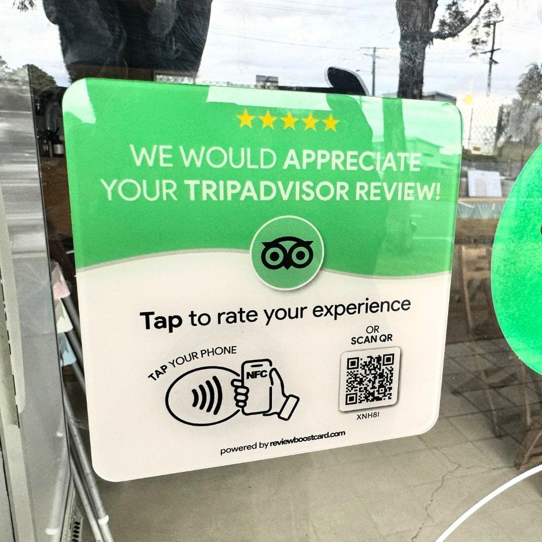 A ReviewBoost card requesting a TripAdvisor review placed on a window. The card is green and white with the TripAdvisor logo and text that says, 'We would appreciate your TripAdvisor review! Tap to rate your experience. Tap your phone or scan QR