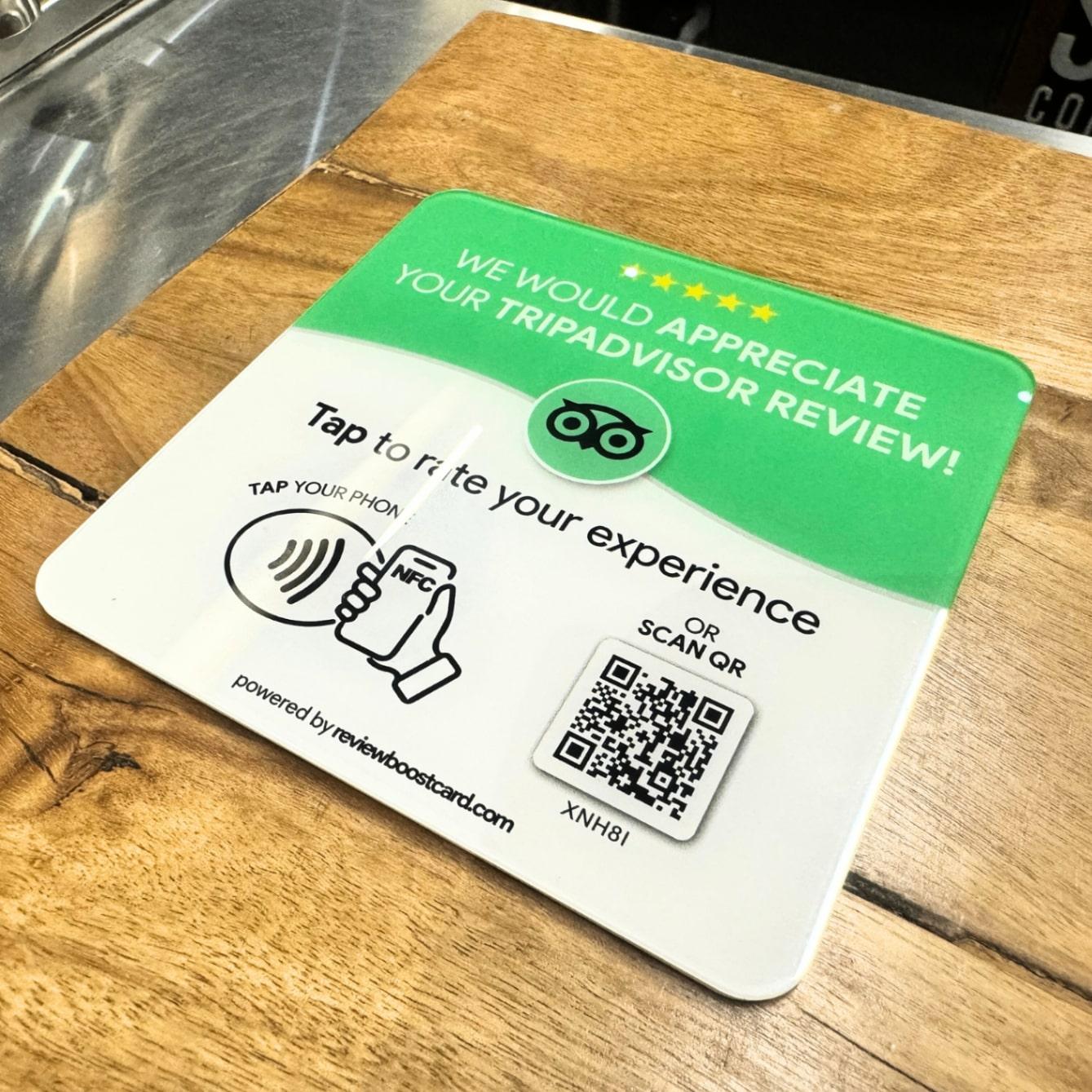 A ReviewBoost card requesting a TripAdvisor review placed on a wooden counter. The card is green and white with the TripAdvisor logo and text that says, 'We would appreciate your TripAdvisor review! Tap to rate your experience. Tap your phone or scan QR