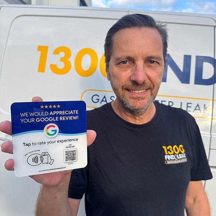 A man holding a ReviewBoost plaque in front of a 1300 Find Leak van, encouraging customers to leave a Google review by tapping or scanning with their phone