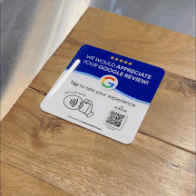GIF showing a ReviewBoost plaque on a wooden table. The plaque reads "We Would Appreciate Your Google Review!" with a QR code and NFC tap area, illustrating the ease of leaving a Google review by simply scanning or tapping